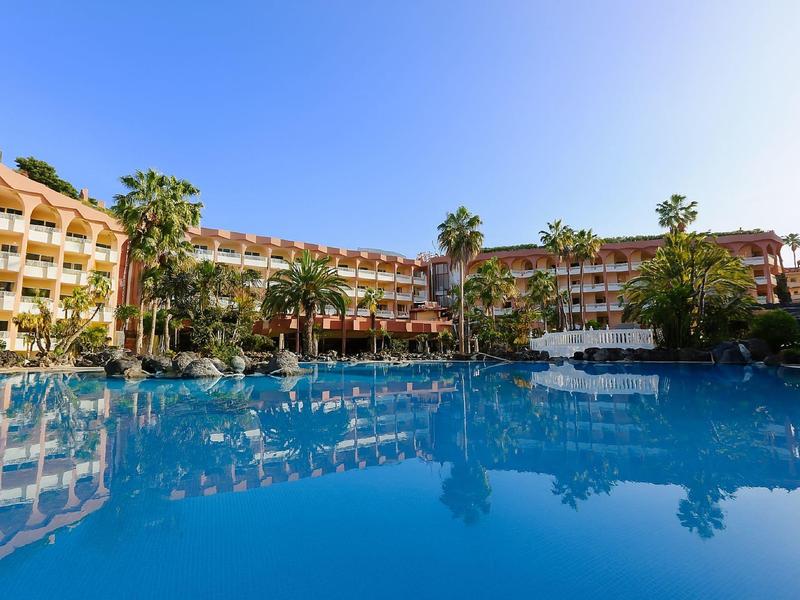 Grande piscina dell'hotel con palme e edificio a più piani sotto un cielo limpido.