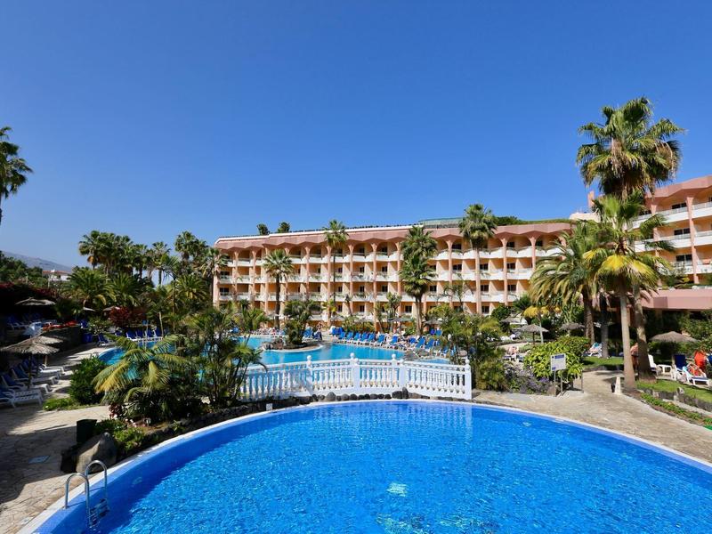 Grande hotel con piscina rotonda blu e ponte, circondato da palme sotto un cielo azzurro chiaro.