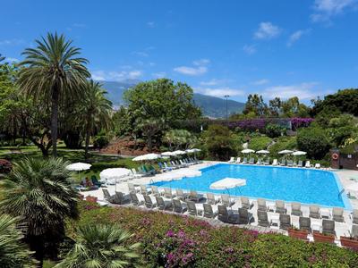 Velký bazén s lehátky a slunečníky v bujné zahradě pod modrou oblohou.