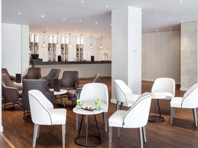 Moderní hotelová lounge se světlými polstrovanými židlemi, malými stolky a barem v pozadí.