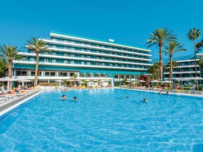 Großer blauer Pool vor modernem Hotel mit Palmen und Sonnenliegen bei klarem Himmel.