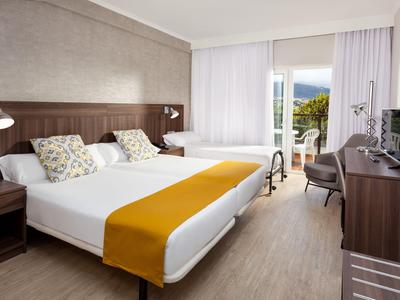 Světý hotelový pokoj se dvěma oddělenými postelemi, žlutými běhouny, psacím stolem a balkonem s výhledem na hory.