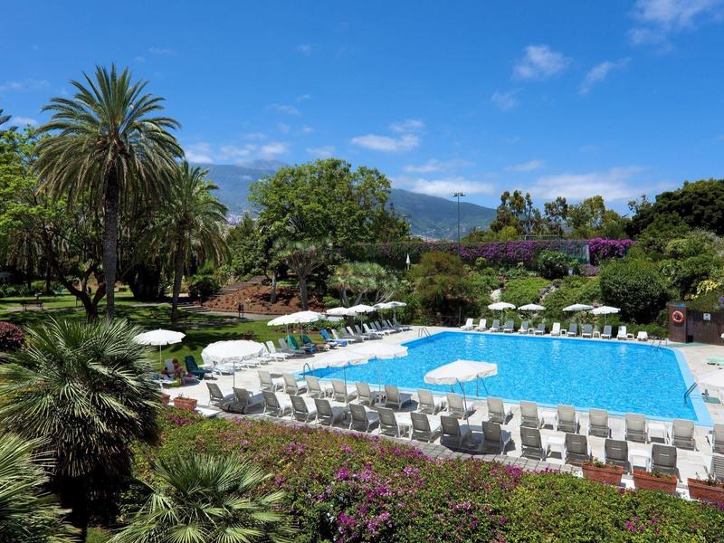 Velký bazén s lehátky a slunečníky v bujné zahradě pod modrou oblohou.