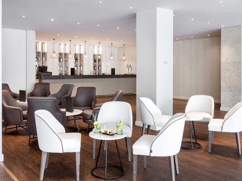 Moderní hotelová lounge se světlými polstrovanými židlemi, malými stolky a barem v pozadí.