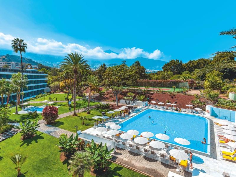 Venku bazén s deštníky a lehátky obklopený palmami a zelenými zahradami.