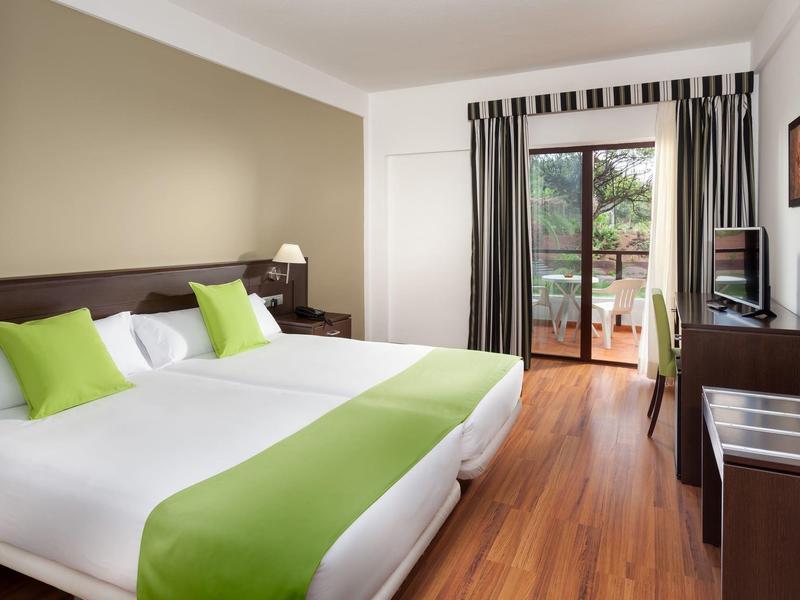 Moderní hotelový pokoj s manželskou postelí, zelenobílým povlečením a balkonem s výhledem ven.