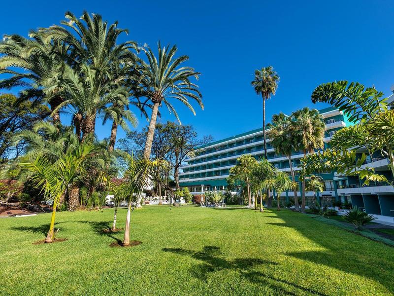 Moderní hotel obklopený zeleným trávníkem a palmami pod jasnou modrou oblohou.