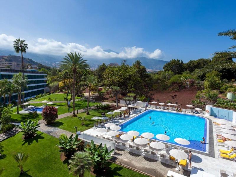 Hotelový bazén s lehátky a palmami, obklopený zelenými zahradami a horami v pozadí.