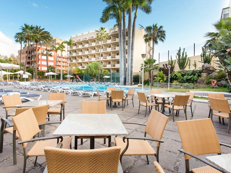 Terraza del hotel con mesas y sillas frente a la piscina y palmeras bajo el cielo azul.
