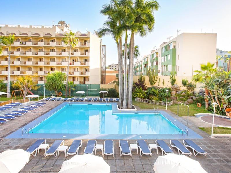 Área de piscina clara con tumbonas, rodeada de edificios hoteleros y palmeras bajo cielo azul.