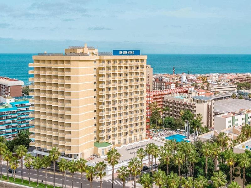 Gran edificio de hotel cerca del mar con piscinas y palmeras verdes en un paisaje urbano.