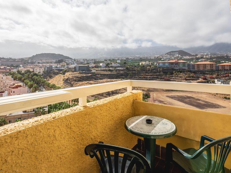 Balcón con mesa pequeña y dos sillas con vista a una ciudad y paisaje montañoso bajo cielo nublado.
