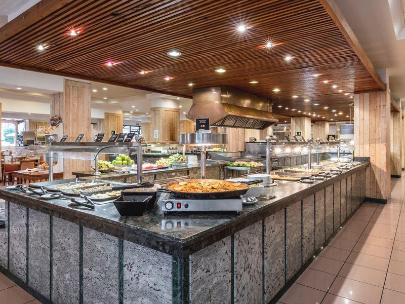 Área de buffet con diversas comidas en restaurante moderno y bien iluminado con techo de madera y suelo de baldosas.