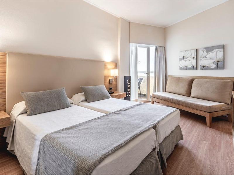 Habitación de hotel luminosa con cama doble, sofá, cuadros en la pared y suelo de madera.