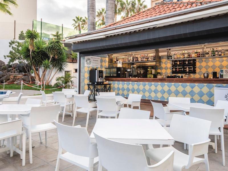 Área exterior moderna de un hotel con mobiliario blanco y bar bajo un área cubierta.