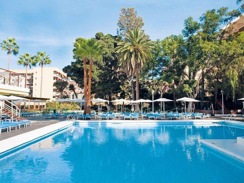 Gran piscina al aire libre con tumbonas y sombrillas, rodeada de palmeras y edificios.