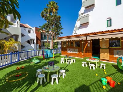 Groene tuin met kinderspeelgoed en tafels naast een wit gebouw bij zonnig weer.