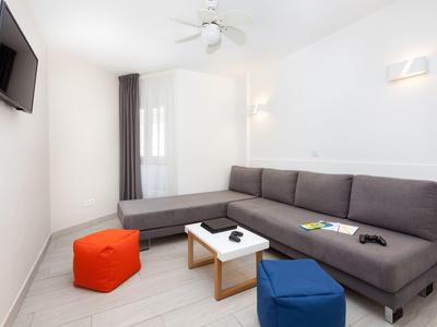 Moderne woonkamer met grijze hoekbank, kleurrijke poefs en wand-tv in lichte kamer.