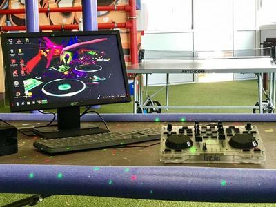 Computer met DJ-software naast een mengpaneel en tafeltennistafel in een kamer met groen tapijt.