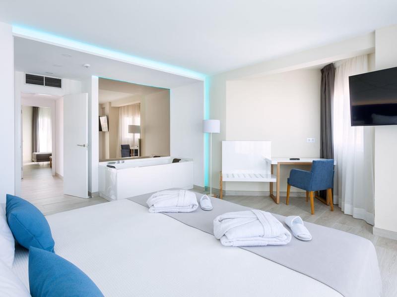 Lichte hotelkamer met tweepersoonsbed, tv en open badkamer met bad.