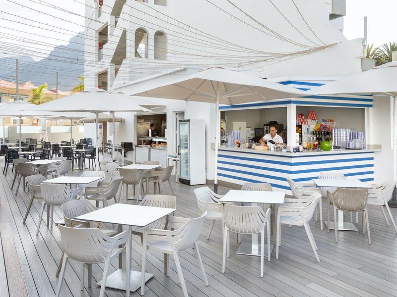 Buitenrestaurant met witte tafels en stoelen en een blauw-wit gestreept snackkraampje.