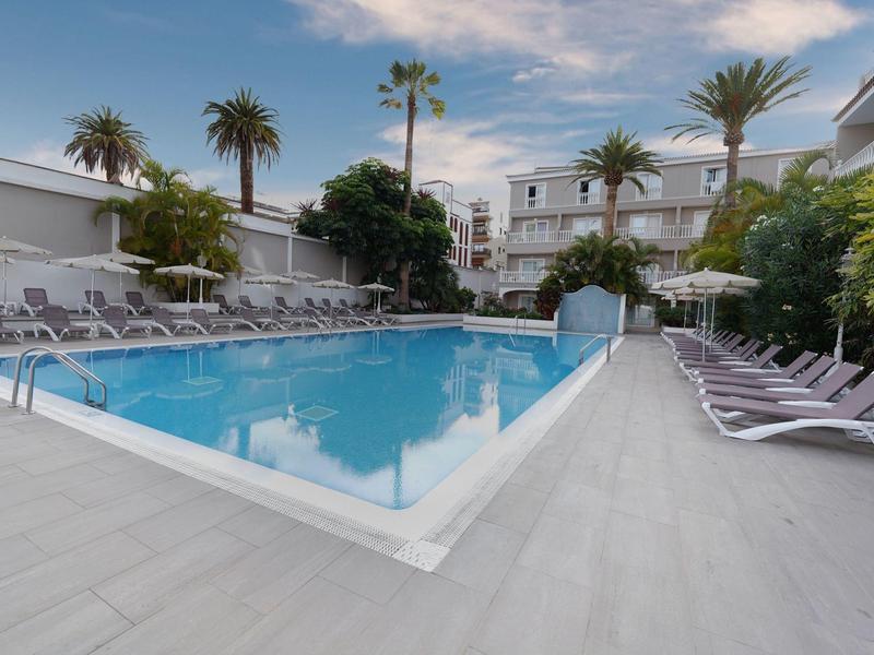 Modern outdoor pool with lounge chairs and palm trees at a hotel surrounded by white buildings.