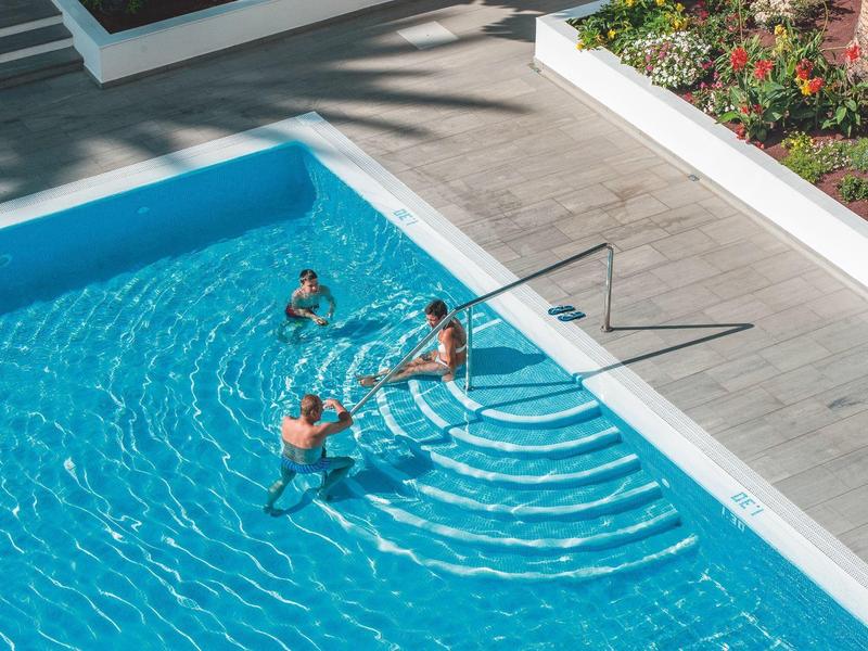 Three people enjoying a sunny swimming pool with clear blue water and wooden deck.