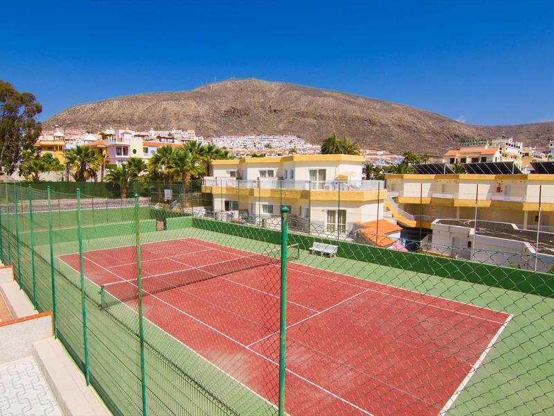 Court de tennis rouge avec clôture verte devant des bâtiments jaunes et un paysage de montagne en arrière-plan.