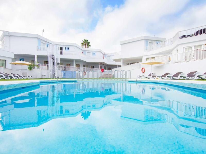 Piscine claire devant des bâtiments d'hôtel blancs avec des transats sous un ciel bleu.