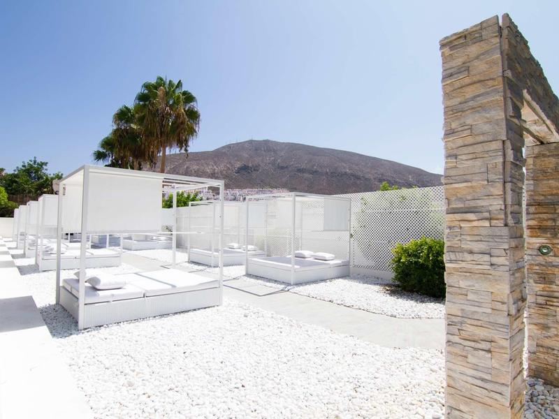 Zone de sable blanc avec des lits modernes couverts de blanc et un pilier en pierre sous un ciel clair.