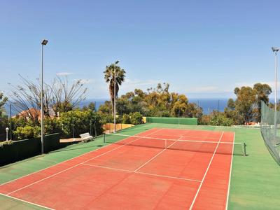 Roter Tennisplatz im Freien mit grünem Rand und Palmen unter klarem Himmel mit Blick auf das Meer.