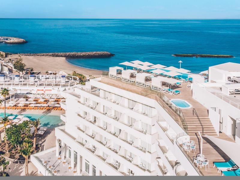 Modern hotelcomplex met zwembaden, witte gebouwen en uitzicht op de blauwe zee en het zandstrand.