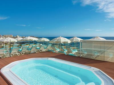 Jacuzzi en una terraza con vista al mar y tumbonas bajo sombrillas en la playa.