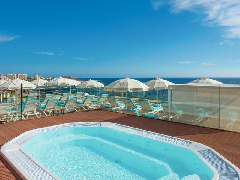 Jacuzzi en una terraza con vista al mar y tumbonas bajo sombrillas en la playa.