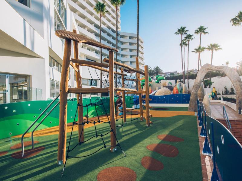 Parque infantil con estructura para trepar y toboganes junto a un hotel al atardecer.