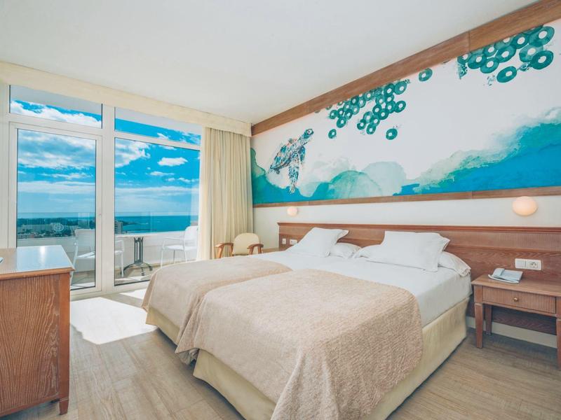 Habitación de hotel luminosa con cama grande y decoración de pared marina junto a ventana con vista al mar.