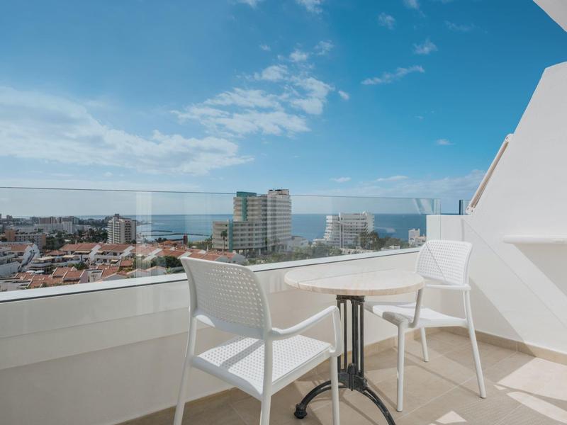 Balcón con dos sillas blancas y mesa, vista a la ciudad y el mar bajo cielo azul.