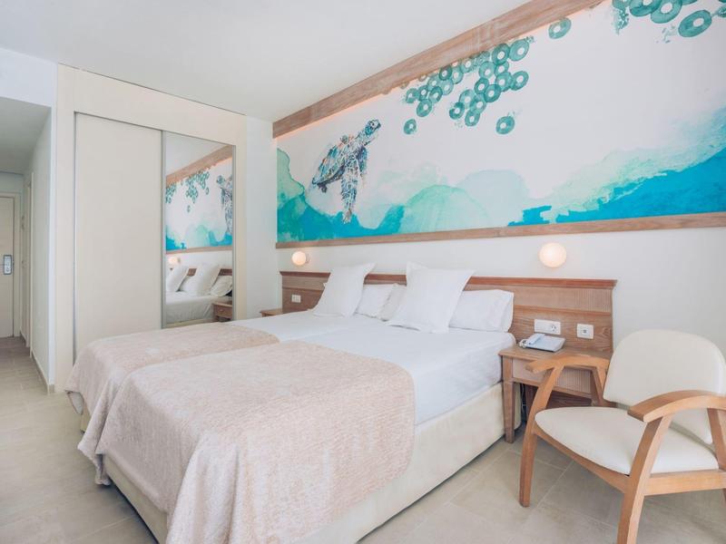 Habitación de hotel luminosa con cama doble, mesita, silla y cuadro marino decorativo sobre la cabecera.