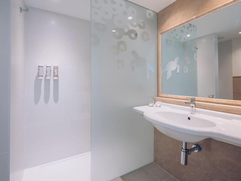 Área moderna de baño de hotel con pared de ducha de vidrio y lavabo doble en diseño claro.