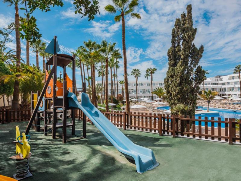 Área de juegos infantil con tobogán junto a piscina y palmeras bajo cielo despejado.