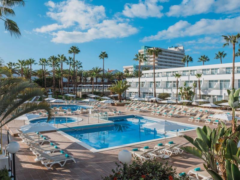 Gran piscina de hotel con tumbonas y palmeras bajo un cielo azul.