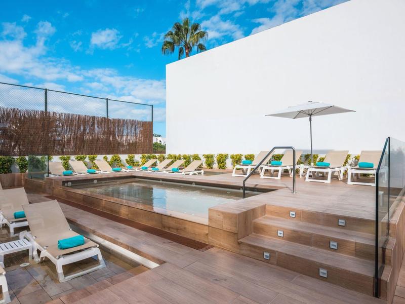 Área moderna de piscina al aire libre con tumbonas y sombrillas junto a una pared blanca bajo el cielo azul.