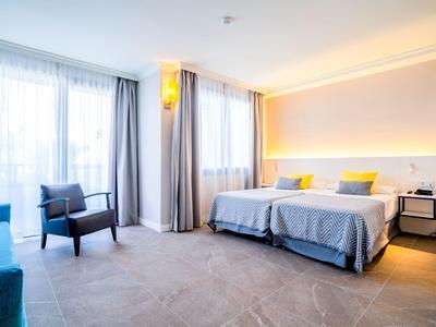 Moderní hotelový pokoj se dvěma postelemi, křeslem, pohovkou a velkými okny se závěsy.