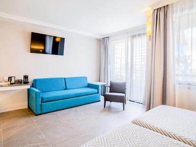 Světle hotelový pokoj s modrou pohovkou, křeslem, televizí na zdi a velkými okny s záclonami.