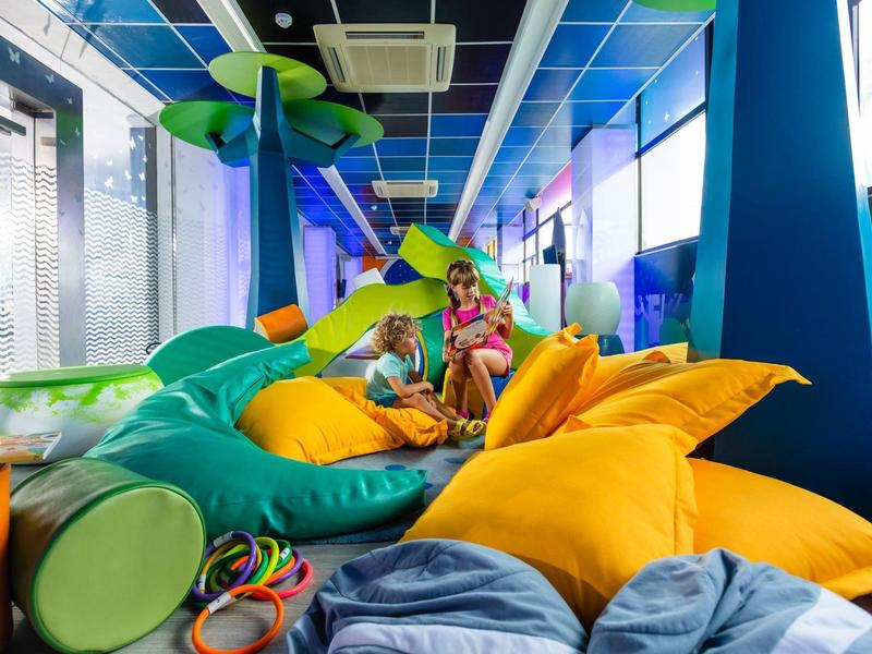 Děti si hrají v barevné vnitřní herně s velkými polštáři a skluzavkou.