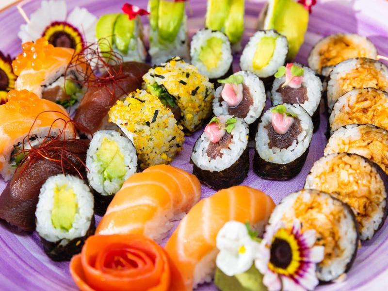 Barevný talíř sushi s různými rolkami a čerstvým lososem, zdobený květinami.