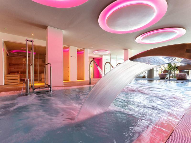 Moderní hotelový bazén s vodopádem a růžově osvětleným stropem.