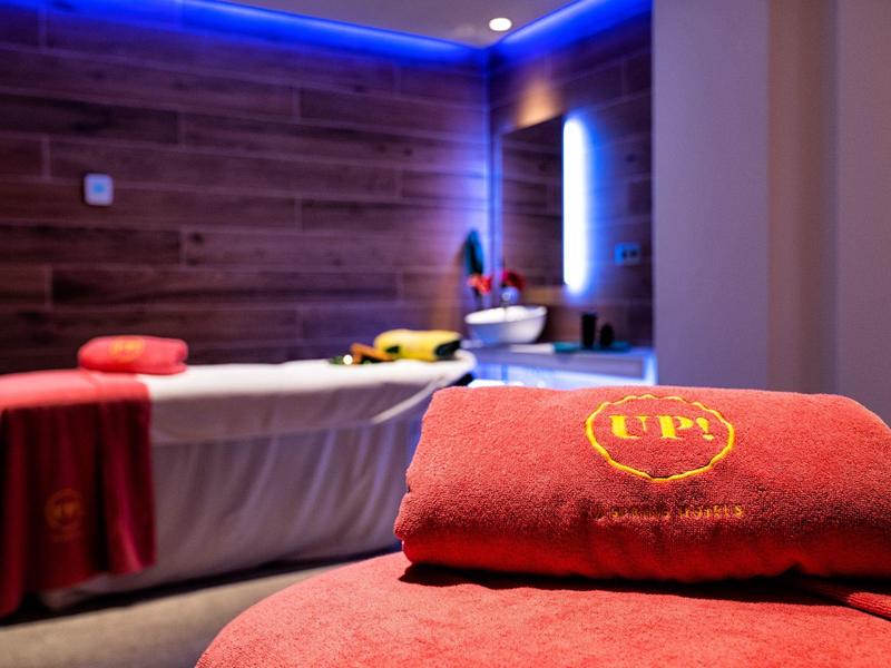 Červený ručník s logem hotelu leží v moderní spa místnosti s masážními lehátky a modrým osvětlením.