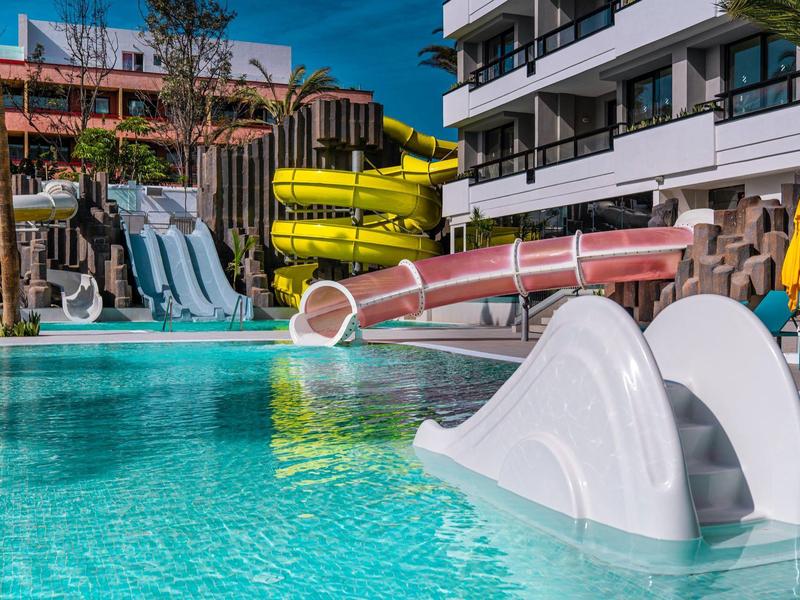 Bazén s vodními skluzavkami a malou skluzavkou v hotelovém komplexu pod modrou oblohou.