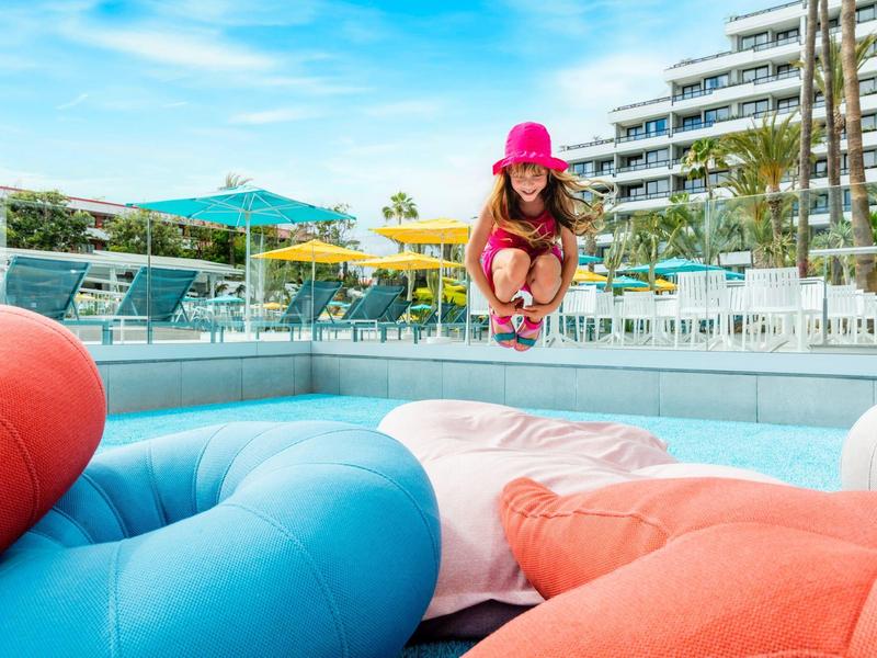 Dítě v červeném klobouku skáče do bazénu s velkými barevnými plovoucími kruhy.